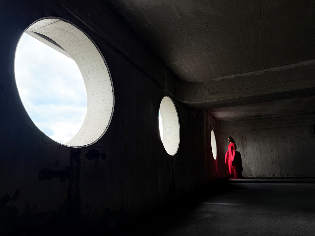 A woman wearing a long red dress stands in an empty room being photographed. The Photo app toggles through the focal lengths to demonstrate the capabilities of the pro camera system, showcasing: 24 mm
